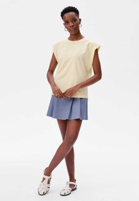 Lichtgele top met korte mouwen en een relaxte pasvorm, gecombineerd met een blauwe geplooide rok en witte sandalen met bandjes, staand op een neutrale achtergrond.