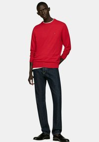 Maglione di cotone rosso con scollo a girocollo, polsini e orlo a coste. Abbinarlo a jeans di denim blu scuro e scarpe nere, mentre si trova in piedi su uno sfondo bianco.