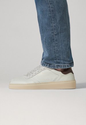 Zapatilla blanca de cuero con suela beige y lengüeta trasera marrón, usada con vaqueros azules contra un fondo claro y liso.