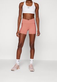 Bílý sportovní podprsenka s černým logem, spárovaná s broskvovými šortkami s vysokým pasem. Model nosí světle růžové sportovní boty a bílé ponožky.