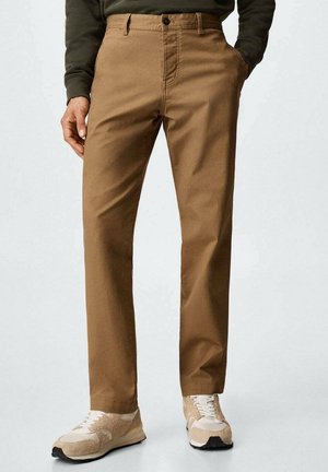 Mand iført brune bukser med lige ben og beige sneakers, hænderne i lommerne, foran en ensfarvet lys baggrund.