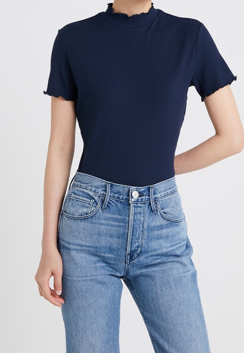 Kobieta w granatowej prążkowanej bluzce z krótkim rękawem wykończonej falbanką, w połączeniu z jasnoniebieskimi dżinsami z wysokim stanem.