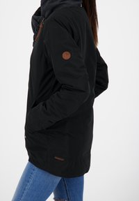 Czarna zewnętrzna kurtka z gładkiego materiału, z kieszenią na lewym rękawie i brązową naszywką z logo. Dopasowana do niebieskich jeansów.