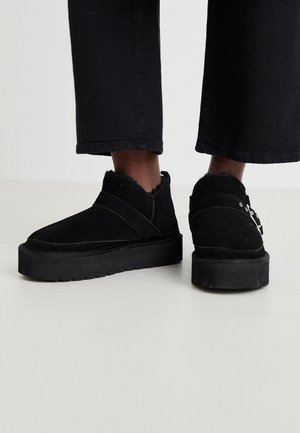 Czarne zamszowe botki na płaskiej podeszwie z platformą, z krzyżującymi się paskami i futrzanym wykończeniem przy kołnierzu.