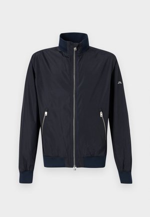 Tamsiai mėlyna lengva striukė su užtrauktuku, aukštu apykakliu, elastinėmis rankogaliais ir apačia, bei užtrauktukais užsegamomis šoninėmis kišenėmis ant vientisos spalvos fono.