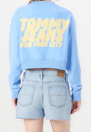 Osoba ubrana w jasnoniebieską krótką bluzę z napisem "TOMMY JEANS NEW YORK CITY" w żółtym kolorze oraz jasne, krótkie spodenki dżinsowe, widok od tyłu.