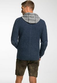 Marineblauwe gebreide trui met grijze capuchon, voorzien van elleboogpatches en geribbelde manchetten. Draag over bruine shorts, die de gestructureerde stof laten zien.