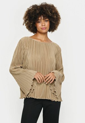 Blusa beige plissettata con ampio scollo, maniche a campana ruffles e finitura testurizzata, abbinata a pantaloni neri.