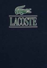 Tamsiai mėlynas medvilninis marškinėliai su žaliu krokodilo logotipu ir baltu tekstu "LACOSTE" horizontaliose linijose po logotipu.