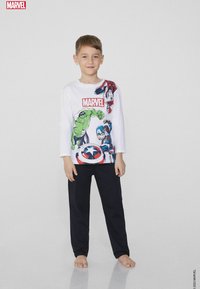 Biała koszula z długim rękawem ozdobiona kolorowymi grafikami superbohaterów, z logo Marvela. W połączeniu z czarnymi spodniami, model stoi boso.