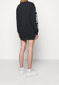 Černé oversized šaty ve stylu mikiny s dlouhými rukávy, s bílým textem na rukávu, vyrobené z měkké látky, s žebrovaným lemem.