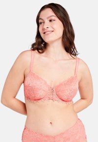 Bralette de encaje en color coral con estampado floral, tirantes ajustables y detalle de centro fruncido. Textura suave y bordes ondulados distintivos.