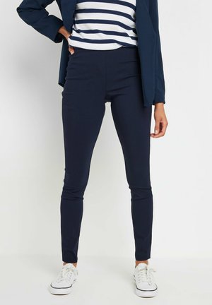 Kvinde iført marineblå tætsiddende bukser, hvide sneakers, en hvid og marineblå stribet top og en marineblå blazer, stående med den ene hånd på hoften.