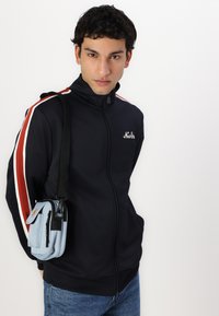 Ναυτικές ζακέτες με μπλε φερμουάρ και κόκκινες και λευκές ρίγες, σε συνδυασμό με μια μικρή ανοιχτό μπλε τσάντα ώμου με φερμουάρ και κλιπ.