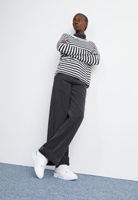 Stripet langermet topp i svart og hvitt, kombinert med løstsittende grå bukser og hvite sneakers, mot en ensfarget hvit bakgrunn.