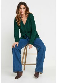Grön kabelstickad cardigan med djup V-ringning och svarta knappar, kombinerad med blå vida jeans och bruna ankelstövlar.