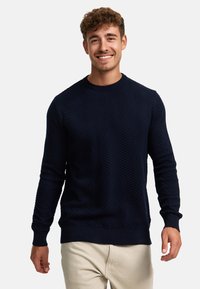 Mørkeblå genser med teksturert chevronmønster, strikket stoff, rund halslinning og ribbestrikket mansjetter og kant. Båret over beige bukser.