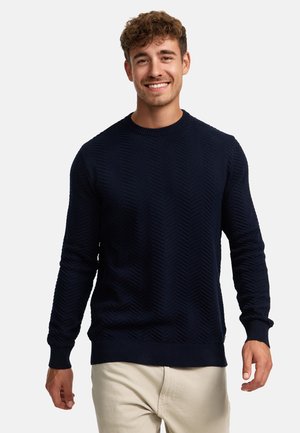 Marineblauwe trui met een getextureerd chevronpatroon, gebreide stof, ronde halslijn en geribde mouwen en onderrand. Draagt over beige broek.