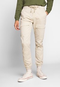 Beige-hii cargo pants, iina elastic cuffs, with adjustable drawstring waist, ama pockets nyingi. Mavazi ya pamoja na white high-top sneakers.