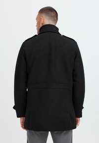 Schwarzer Wollmantel mit hohem Kragen, Schulterklappen und Seitentaschen. Er hat einen taillierten Schnitt und eine glatte Textur.
