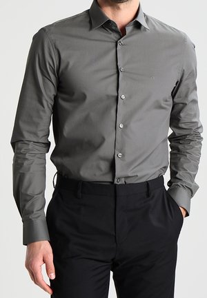 Hombre con camisa ajustada de botones de color gris oscuro y mangas largas, y pantalones de vestir negros, de pie con una mano en el bolsillo.