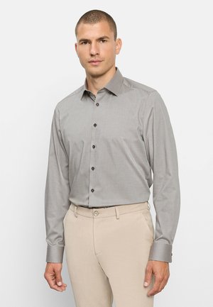 Man draagt een lichtgrijs geruit overhemd met lange mouwen en beige broek, staand tegen een effen witte achtergrond.
