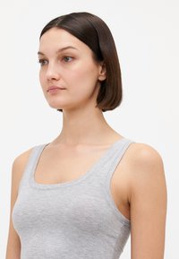 Jeune femme aux cheveux bruns courts portant un débardeur gris clair, tournée vers la gauche sur un fond blanc uni.