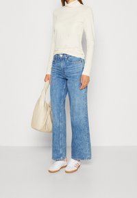 Lichtgewicht crème coltrui, blauwe wijde jeans, witte sneakers met roze accenten en een crème handtas, tegen een neutrale achtergrond.