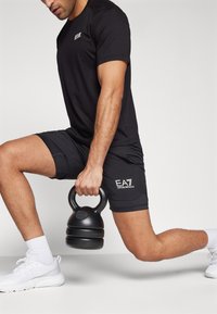 Juodas sportinis marškinėlis ir šortai, laikantis juodą kettlebell. Balti sportbačiai su išsiskiriančiu logotipu ant šortų. Švarus dizainas su paprastomis linijomis.
