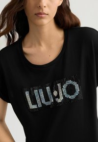 T-shirt noir à manches courtes avec un logo brillant et texturé portant l'inscription "LIU JO" en lettres réfléchissantes sur le devant.