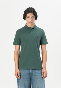 Polo shirt vert foncé à manches courtes, avec un col et un logo noir sur la poitrine gauche. Design minimaliste, texture douce.