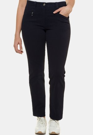 Femme portant un pantalon noir cintré avec des poches avant zippées et des baskets blanches, main reposant dans la poche droite sur fond uni.
