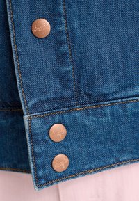 Džinsinio švarko rankogalis su trimis apvaliais vario spalvos mygtukais, pasižymintis siūtu siūleliu ir ryškiai mėlyna medžiaga. Džinse matyti subtili tekstūra.