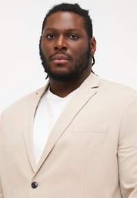 Beige suit jacket with a single button closure, lapel collar, and a chest pocket, worn over a white t-shirt. Smooth fabric texture.