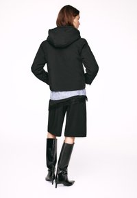 Crna skraćena jakna s kapuljačom, nošena preko plave majice s čipkastim rubom, u kombinaciji s crnim štiklama do koljena i crnim kratkim hlačama.