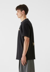 Melnas kokvilnas t-krekls ar īsām piedurknēm, apaļu kakla izgriezumu, ar zeltītu tekstu uz priekšpuses un aizmugures. Tieša griezuma, plaša piegriezuma, gluda tekstūra.