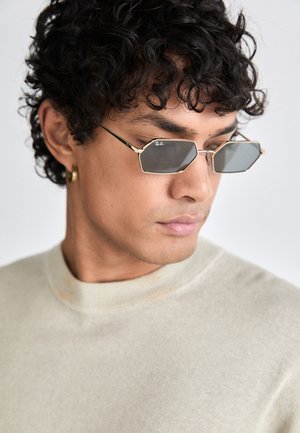 Hexagonale zonnebril met een gouden metalen frame en donkere lenzen. Draagt een lichtbeige gebreide shirt met een ronde hals.