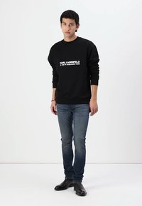 Crna dukserica s bijelim printanim tekstom, plave traperice i crne cipele. Dukserica ima okrugli izrez i duge rukave.