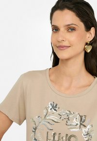 T-shirt beige à manches courtes avec un col rond, orné d'un motif floral en sequins argentés et noirs. Matière en coton doux, coupe décontractée. Boucles d'oreilles en forme de cœur doré.