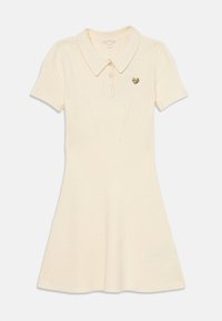 Korte ermed polo kjole i kremfarge, ribbet tekstur, fitted overdelen, utflared skjørt, har krage og et lite gyllent emblem på brystet.