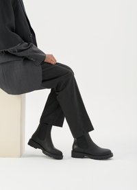 Melnas ādas potītes zābaki ar elastīgām sānu malām, masīvu gumijas zoli un teksturētu apdari. Vilkti ar oglekļa pelēkiem biksēm un žaketi.