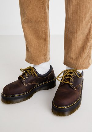 Braune Lederschuhe mit gelben Nähten, runder Spitze, dickem Plateausohle und kontrastierenden schwarzen Akzenten. Getragen mit weißen Socken und Cordhosen.