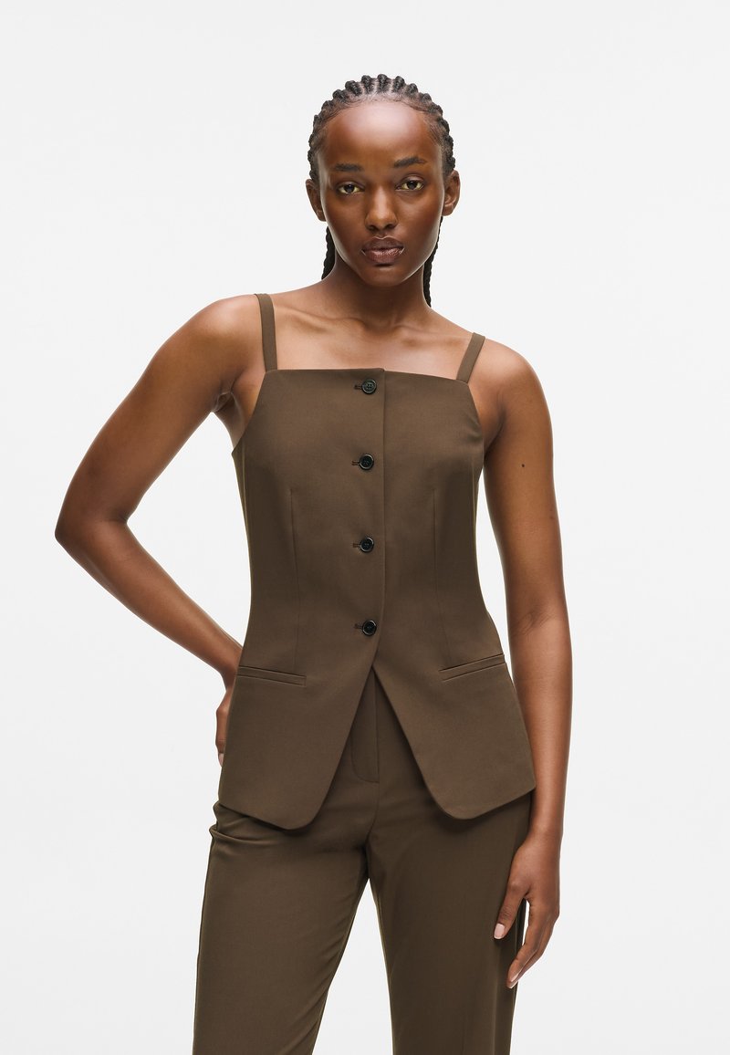 Woman with braided hair wears brown buttoned sleeveless top and matching pants, standing with one hand on hip against white background.