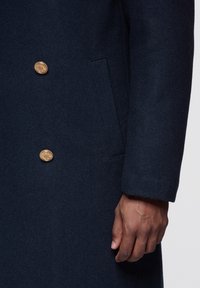 Marineblå uldfrakke med tekstureret finish, dobbeltradet design, med guldfarvede knapper og en sidelomme.