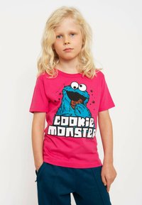 Pink short-sleeve T-shirt featuring a blue Cookie Monster graphic and bold "COOKIE MONSTER" text. Soft cotton material, relaxed fit.