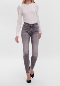 Biała, fakturowana bluzka z długim rękawem połączona z szarymi, obcisłymi dżinsami o wysokim stanie i postrzępionych nogawkach; modelka nosi czarne szpilki.