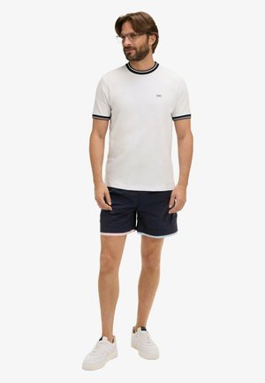 Homme debout portant un t-shirt blanc avec une bordure noire, un short bleu marine avec des bords roses et bleu clair, des baskets blanches et des lunettes.