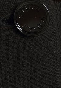 Black knit fabric with a textured surface. Features a prominent round black button embossed with "STEFFEN SCHRAUT."