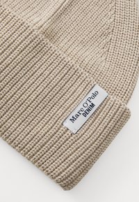 Beige kudumine müts ribakujulise tekstuuriga, millel on valge silt, kus on kirjas "Marc O'Polo DENIM". Materjal näeb välja pehme ja veniv.