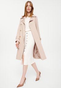 Jasno beżowy trencz z złotymi guzikami, noszony na białej dopasowanej sukience z guzikami z przodu. Modelka ma na sobie brązowe szpilki.